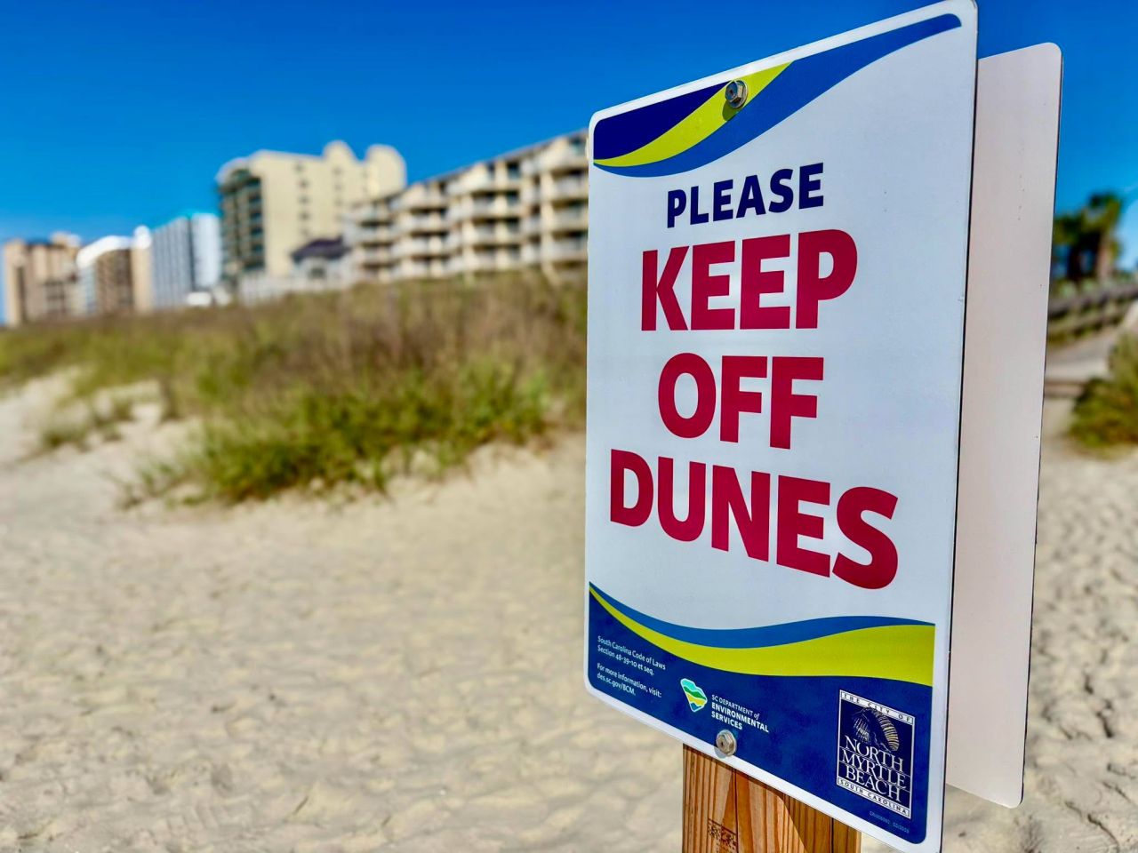 Sign at a beach entrance reading ‘Please Keep Off Dunes,’ with sand, dune grasses, and beachfront buildings in the background under a clear blue sky.