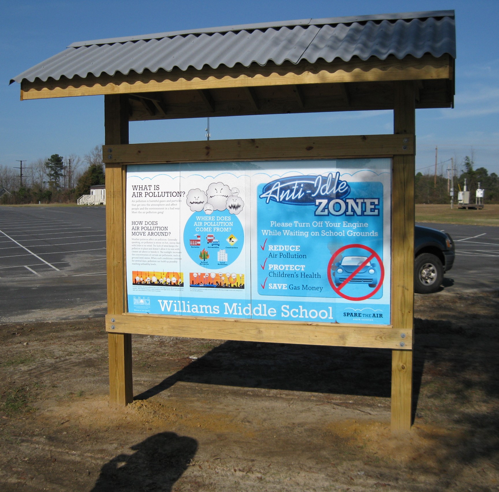 Here’s brief alt text for the image:  “Outdoor wooden bulletin board displaying posters about air pollution and an anti-idling zone at Williams Middle School in a parking lot.”