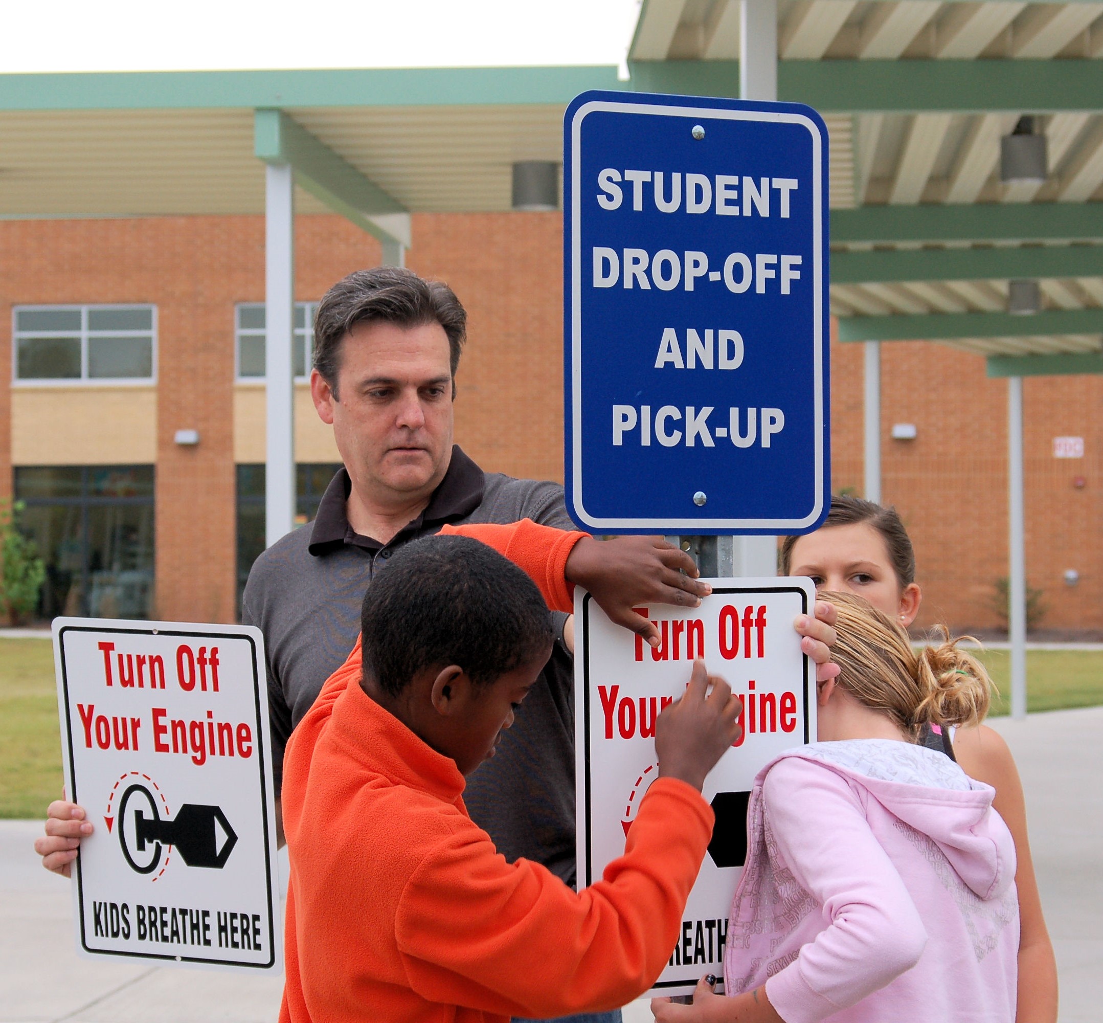 Adults and children stand near a student drop‑off and pick‑up sign, holding signs that read ‘Turn Off Your Engine – Kids Breathe Here’ outside a school building.