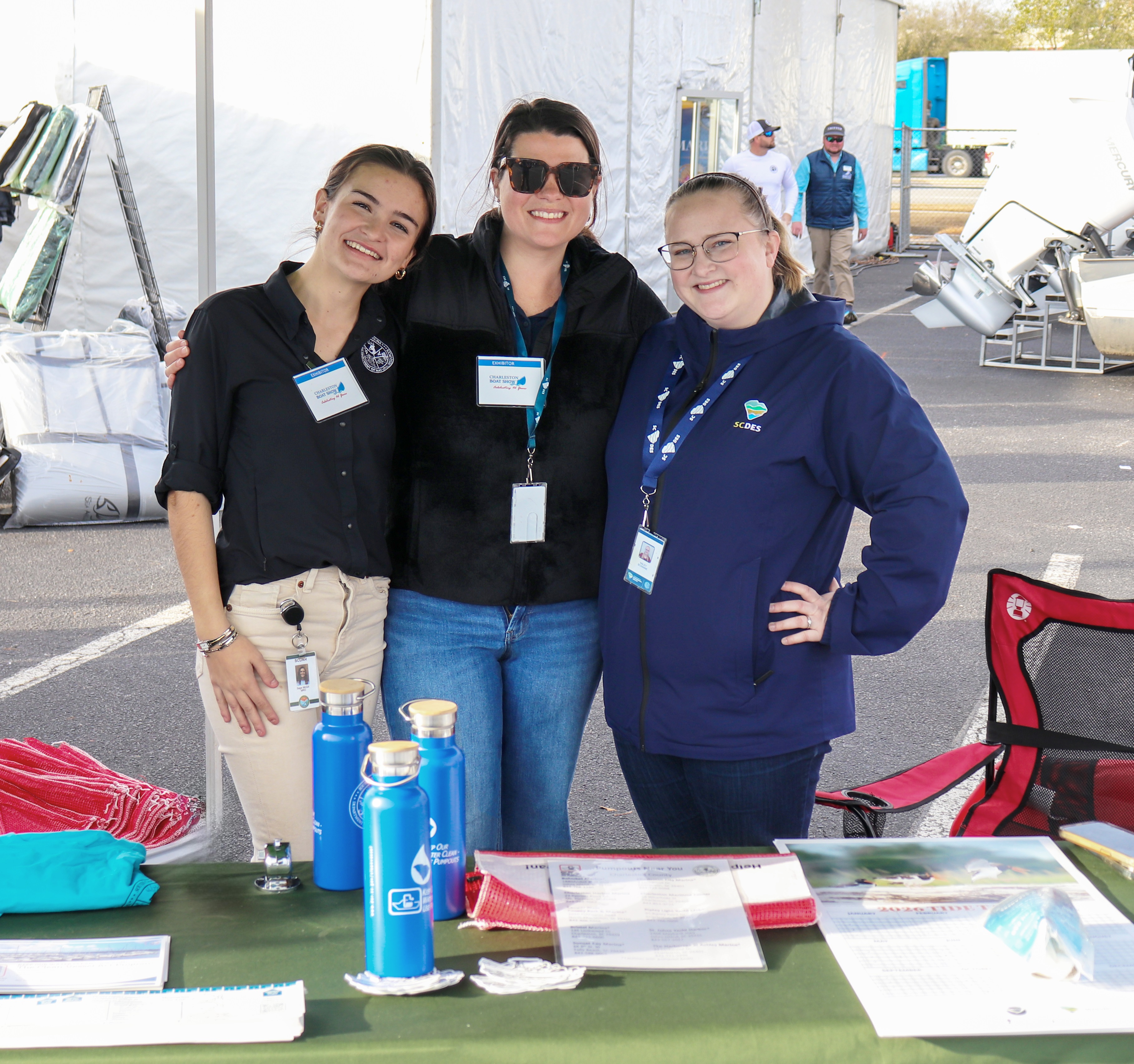 Three people stand together smiling at an outdoor event booth. The booth has informational materials and blue water bottles on the table in front of them. They are wearing lanyards with badges. Tents and event equipment are visible in the background.