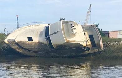 A capsized boat rests partially submerged near a grassy shoreline. Cranes and industrial structures are visible in the background under a clear sky.