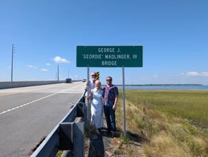 three people stand beside a bridge under a sign reading "George J. 'Geordie' Wallinger, III Bridge"