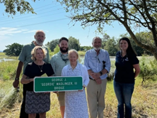 six people stand beside a marsh holding a sign that reads sign reading "George J. 'Geordie' Wallinger, III Bridge"