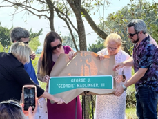 four people unwrap a sign that reads sign reading "George J. 'Geordie' Wallinger, III Bridge" while someone captures photos with an iPhone