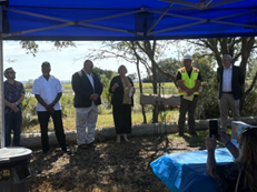 six people stand underneath a blue pop up tent while one woman makes remarks