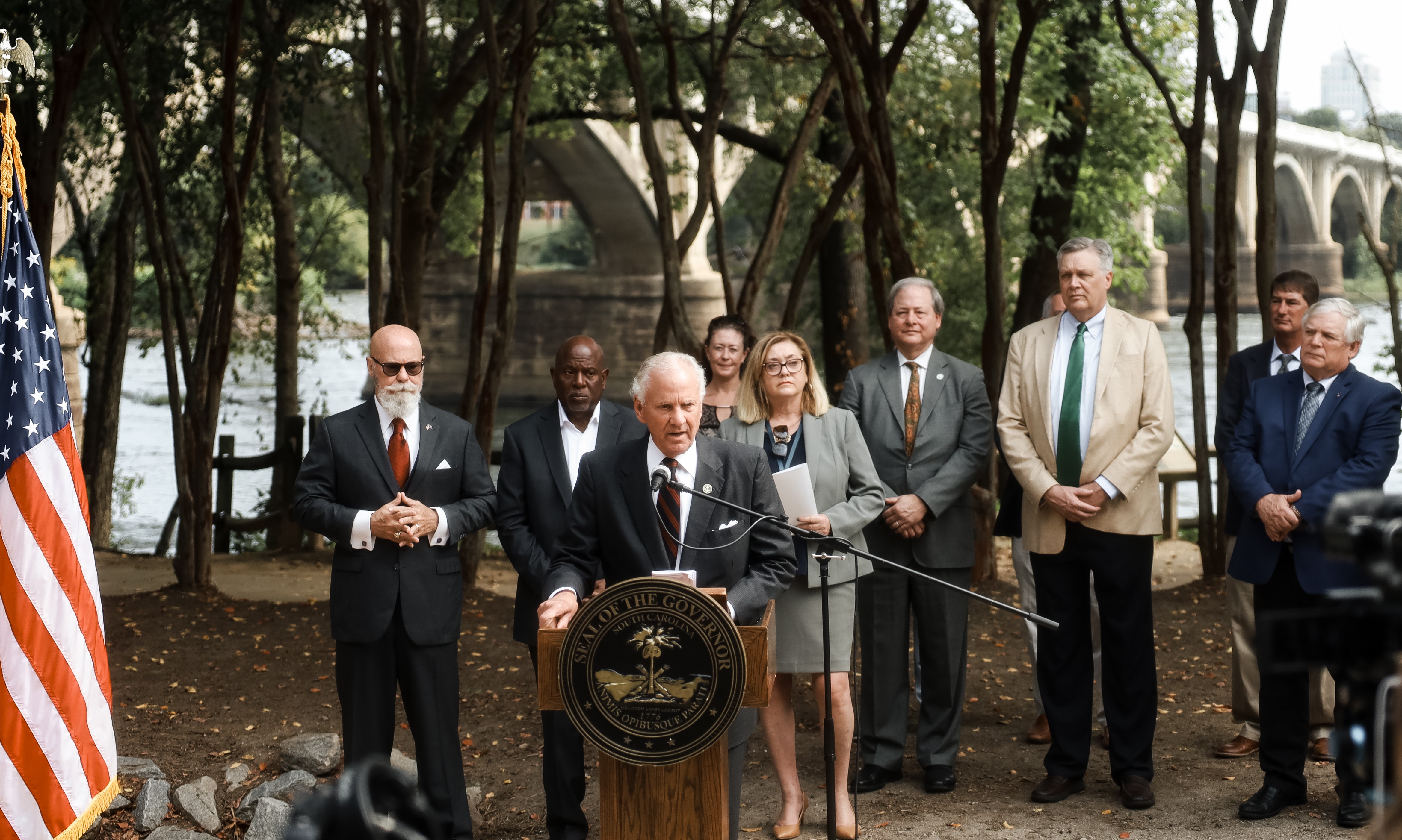 a group of officials stands outdoors by a river with a bridge visible in the background. SC Governor Henry McMaster stands speaking at the podium. 