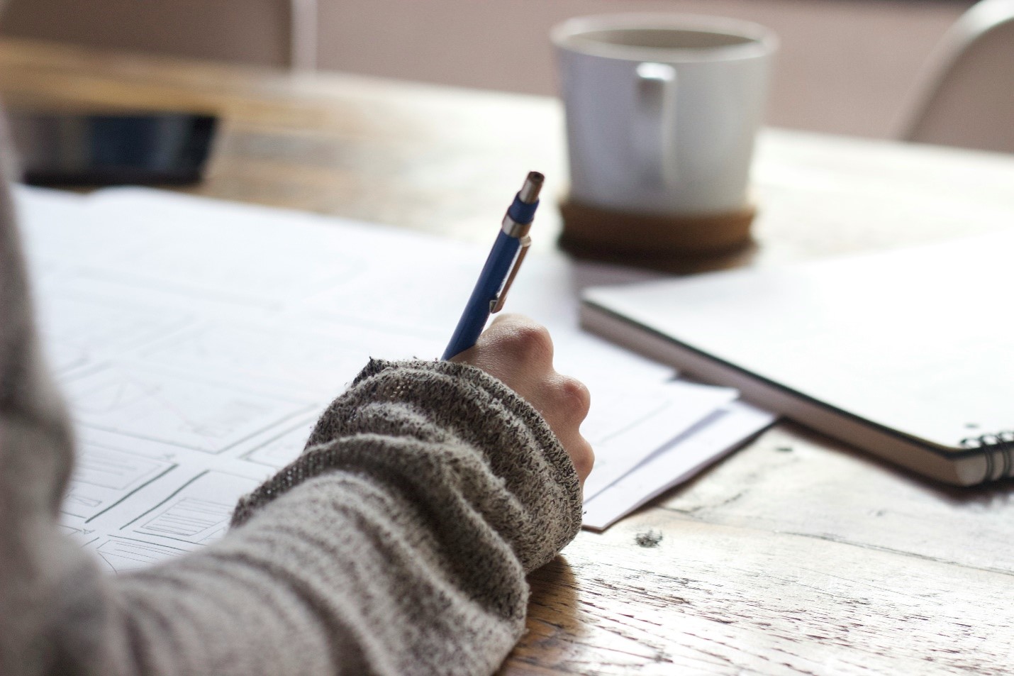 A person’s hand holding a pen and writing on papers spread out on a wooden table. A notebook and a coffee mug sit nearby in a softly lit workspace