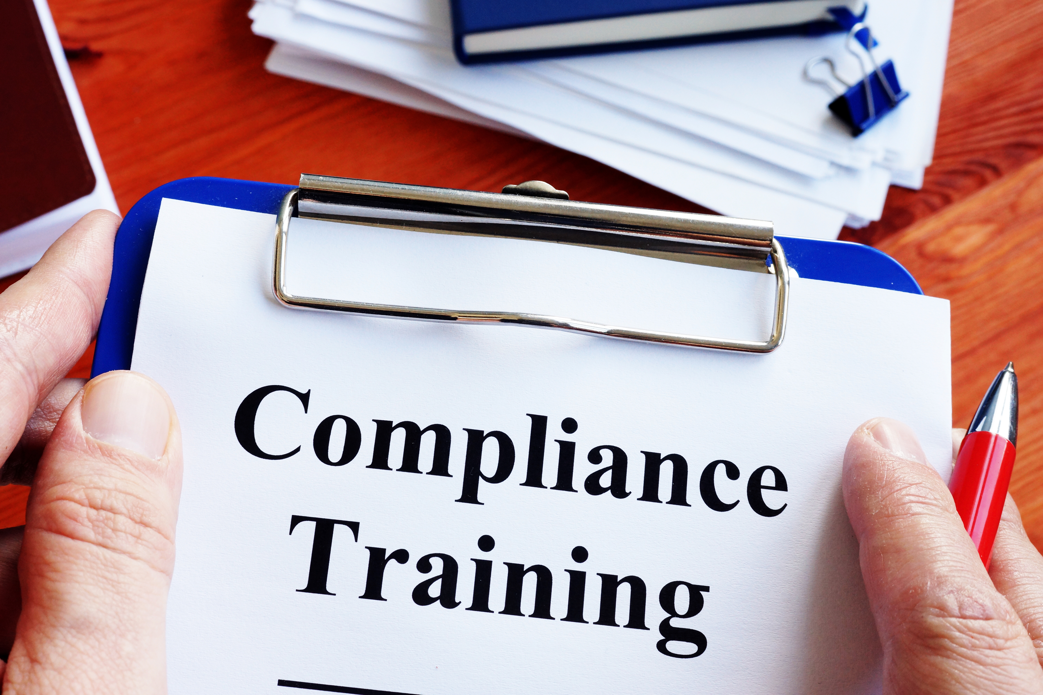 Person holding a clipboard with a paper titled "Compliance Training" and a pen, over a desk with books and documents.