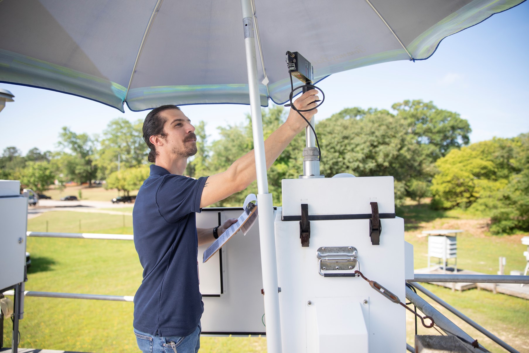 A person is adjusting weather monitoring equipment under a large umbrella, surrounded by greenery.