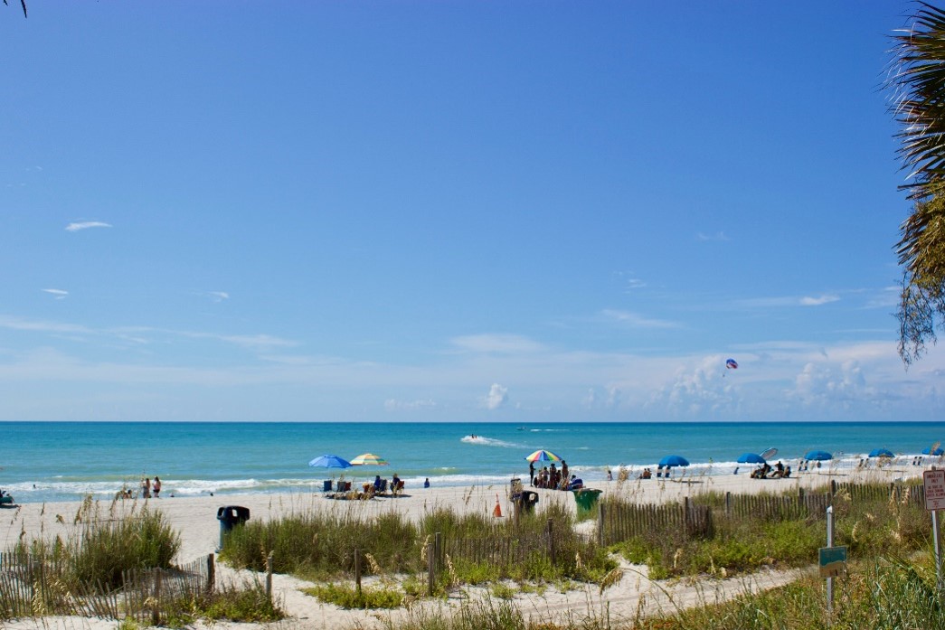 A scenic beach view with a clear blue sky and bright turquoise ocean waves. The sandy beach is lined with grassy dunes and scattered with people relaxing under colorful umbrellas and tents.