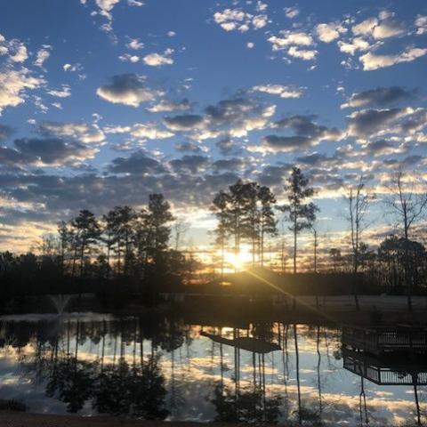 Sunset over a small lake with tall trees silhouetted against the sky. Clouds reflect on the calm water, and a wooden structure with railings is visible along the shoreline.