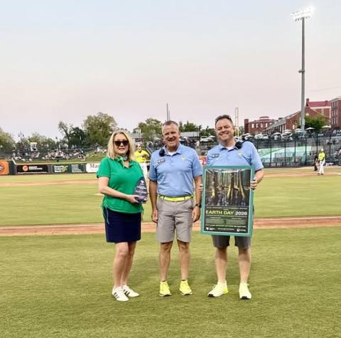 Three individuals standing on a baseball field near the infield. One person is holding an award, and another is holding a poster that reads “Earth Day 2026” with an image of a forest. Stadium seating, players, and field lights are visible in the background.