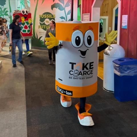 A person in a large battery‑themed costume labeled “Take Charge – Be Battery Smart” walks inside a public exhibit area. The costume has cartoon-style eyes and yellow gloves. In the background, another person is wearing a recycling bin‑themed costume, and several people are walking near colorful displays and signage.
